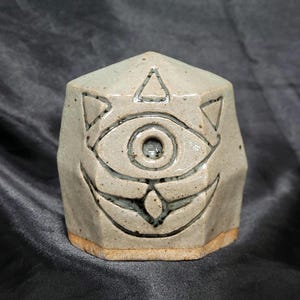 May include: A white ceramic sculpture of a face with a single eye and geometric patterns. The eye is a hole in the sculpture.