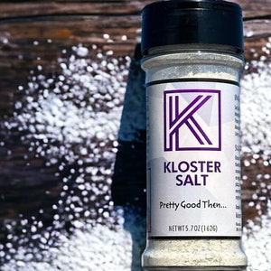 May include: A white plastic jar with a black lid containing salt. The label is white with a purple K logo and the text "Kloster Salt" and "Pretty Good Then..."  The jar is sitting on a wooden surface with white salt scattered around it.  Net Wt 5.7 oz (162g).
