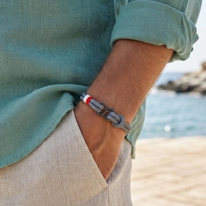May include: A grey cord bracelet featuring a dark metal hook clasp and a small decorative band with red, white, and blue stripes. The bracelet is constructed from two strands of grey cord that are knotted together to form a secure, adjustable loop.