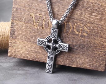 May include: A silver-toned metal Celtic cross pendant featuring intricate knotwork patterns and a central circular design, suspended from a matching wheat chain. The pendant is positioned against a wooden block engraved with the word VIKINGS.