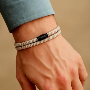 Könnte beinhalten: Ein beiges Seilarmband mit schwarzem Magnetverschluss wird am Handgelenk getragen. Das Armband besteht aus zwei Strängen gedrehten Seils. Der Hintergrund ist einfarbig beige. Die Person trägt ein hellblaues Hemd.