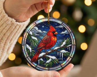 May include: A round, stained-glass style ornament featuring a vibrant red cardinal perched on a snow-covered branch. The background depicts a starry night sky with snow-covered pine needles. The ornament is held by a gold string.