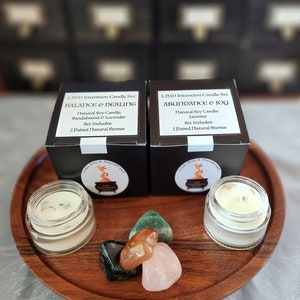 May include: Two black boxes with white text, labeled "LIND Intention Candle Set" and featuring the names "Balance & Healing" and "Abundance & Joy". The boxes contain natural soy candles with corresponding scents and two paired natural stones. The candles are displayed on a wooden tray with four crystals.