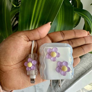 May include: A white phone charger and cable with purple flower accents. The charger has two purple flowers with yellow centers. The cable has a single flower accent. The items are held in a hand.