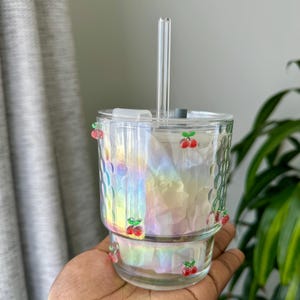 Peut inclure: Un gobelet en verre transparent avec un motif de cerises et une paille en verre transparent. Le gobelet a une finition irisée et un motif texturé. Les cerises sont rouges avec des tiges vertes. Le gobelet est tenu dans une main.