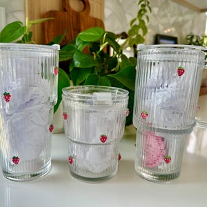 May include: Three clear glass tumblers with vertical ribbed textures and small red strawberry decals. Two tumblers are tall with lids, and one is a shorter, handleless glass. Each piece features a stepped design and contains white paper filler.