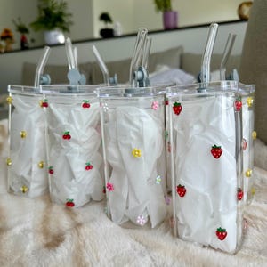 May include: Four clear plastic tumblers with straws, decorated with red strawberries and colorful flower embellishments. Each tumbler contains white tissue paper. The tumblers are sitting on a fluffy, white surface.
