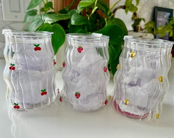 Könnte beinhalten: Drei klare, wellenförmig strukturierte Gläser mit Deckeln. Das erste Glas ist mit roten Kirschmotiven verziert, das zweite mit roten Erdbeermotiven und das dritte mit kleinen gelben Bienenmotiven. Jedes Glas ist mit weißem Seidenpapier gefüllt.