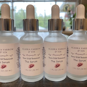 May include: Four frosted glass serum bottles with white droppers and silver collars. Each bottle has a label with the text "ALASKA YARROW & YIN", "Born Wild Botanicals", and "The Serum". The label also includes a rose graphic and states "1 FL OZ (30 ML)".