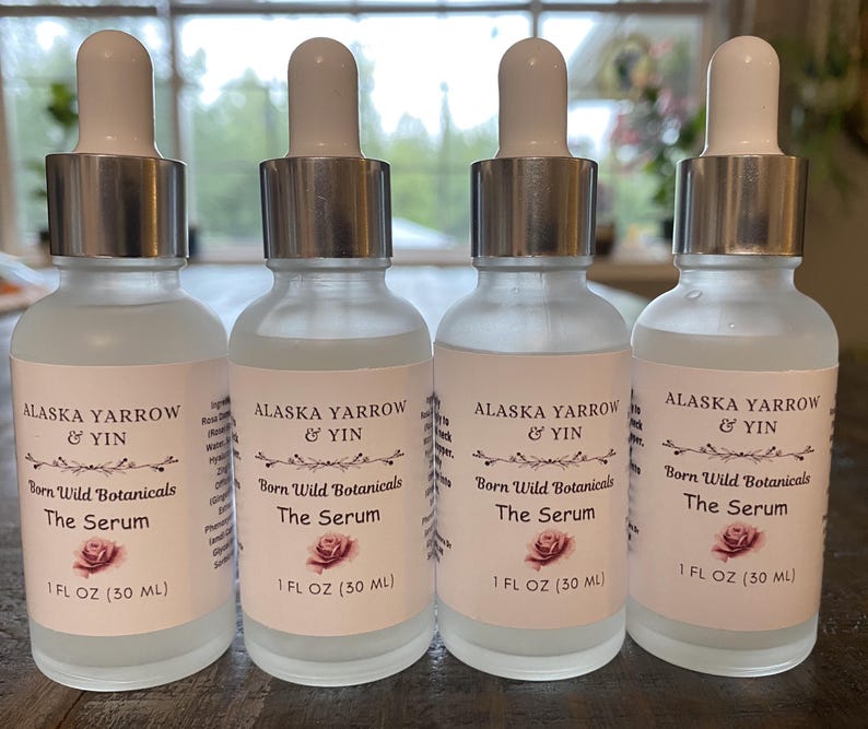 Puede incluir: Cuatro botellas de suero de vidrio esmerilado con cuentagotas blancos y collares plateados. Cada botella tiene una etiqueta con el texto "ALASKA YARROW & YIN", "Born Wild Botanicals" y "The Serum". La etiqueta tambi&eacute;n incluye un gr&aacute;fico de rosa e indica "1 FL OZ (30 ml)".