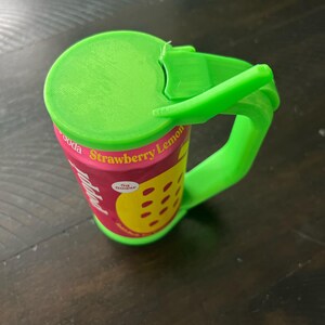 May include: A green 3D-printed soda can with a handle and lid. The can features a pink label with the words "Strawberry Lemon" and a yellow lemon graphic. The handle and lid are bright green.