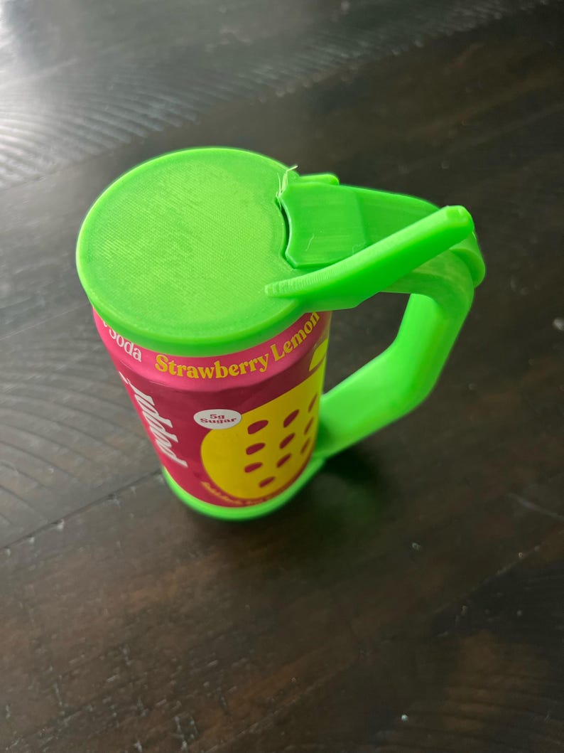 May include: A green 3D-printed soda can with a handle and lid. The can features a pink label with the words "Strawberry Lemon" and a yellow lemon graphic. The handle and lid are bright green.