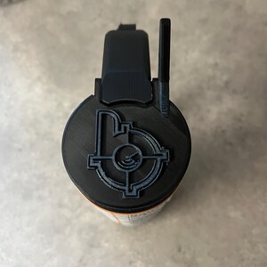 May include: A black and orange cylindrical object with a black top featuring a target design. The top has a handle and a raised sight. The object appears to be 3D printed.