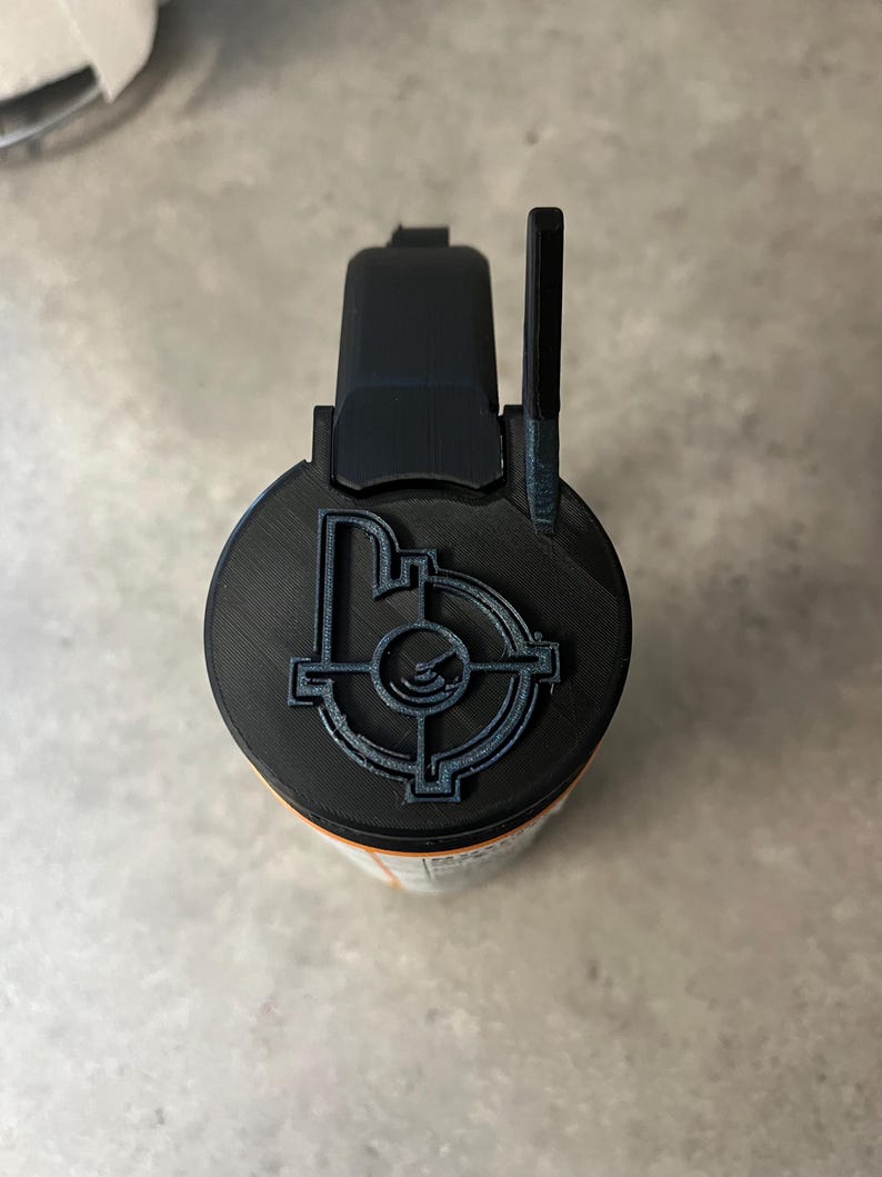 May include: A black and orange cylindrical object with a black top featuring a target design. The top has a handle and a raised sight. The object appears to be 3D printed.