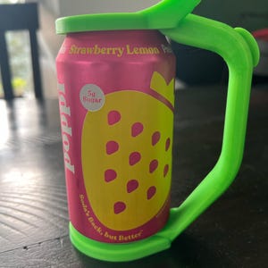 May include: A pink can of "Poppi" soda with a yellow strawberry graphic and the text "Strawberry Lemon Pre". A bright green handle and lid are attached to the can. The can also has the text "Soda's Back, but Better".
