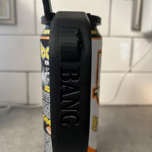 May include: A black 3D-printed can holder with the word "BANG" embossed on the side. The holder is attached to a can of energy drink with orange, yellow, and white labels. The holder has a handle and a top piece that appears to be a bottle opener.