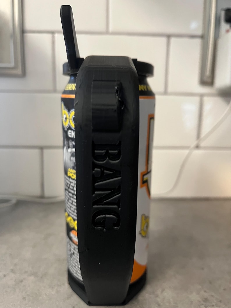 May include: A black 3D-printed can holder with the word "BANG" embossed on the side. The holder is attached to a can of energy drink with orange, yellow, and white labels. The holder has a handle and a top piece that appears to be a bottle opener.
