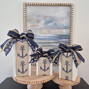 May include: Three decorative wooden boxes wrapped in burlap fabric with navy blue ribbon and anchor print. The boxes are arranged in a descending size order on a wooden tiered stand.