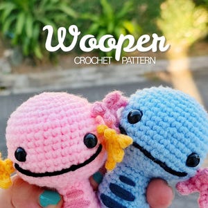 May include: Two crocheted axolotl plushies, one pink and one blue, are held in a person's hands. The axolotls have black eyes, smiling mouths, and yellow and pink frills. The image includes the text "Wooper CROCHET PATTERN".