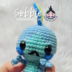 Può includere: Un peluche Sobble all'uncinetto fatto a mano, nei toni del blu e del verde acqua, con grandi occhi neri e piccoli baffi. Il peluche è tenuto in mano e presenta il testo "Sobble Crochet Pattern" e "Abubot ni Kitkat".