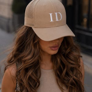 Op de afbeelding: Beige baseballpet met een mesh achterkant en een gebogen klep. De pet heeft de witte letters "ID" op de voorkant. De pet is gemaakt van een zacht materiaal en heeft een klassiek trucker-ontwerp.