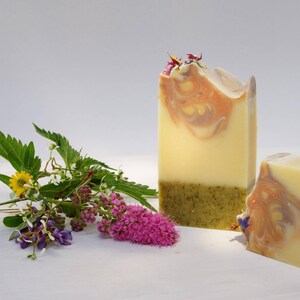 May include: Two rectangular bars of handmade soap with a layered design. The top layer has a swirl of orange and brown, the middle is a pale yellow, and the bottom is green. A sprig of flowers and leaves is next to the soap.