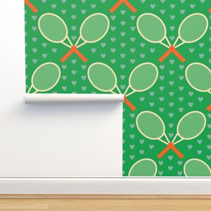 May include: Green wallpaper featuring a repeating pattern of tennis rackets and small gray hearts. The rackets are cream-colored with green netting and orange handles. The wallpaper is partially rolled out, revealing a white backing.