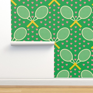 May include: Green wallpaper with a repeating pattern of tennis rackets and pink hearts. The rackets are cream-colored with green netting, and the hearts are evenly spaced across the green background. A roll of the wallpaper is visible.