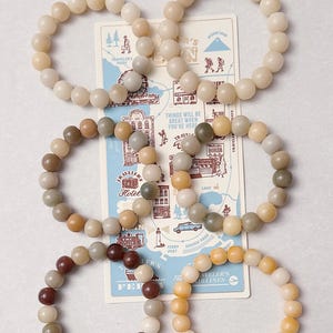 May include: Several beaded bracelets in shades of beige, brown, and yellow are displayed with a vintage-style travel map. The bracelets are made of round beads. The map has illustrations of landmarks and the text "Things will be great when you're here."