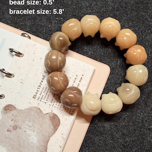 May include: A bracelet featuring rabbit-shaped beads in shades of beige and brown. The bracelet is approximately 5.8 inches in circumference, with individual beads measuring 0.5 inches. The bracelet is displayed on a notebook.