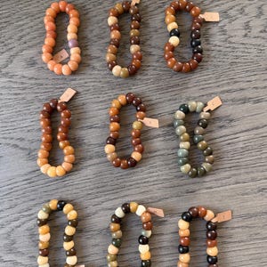May include: Nine beaded bracelets in various colors, including orange, brown, and green. Each bracelet is made of rounded beads and has a small tag attached. The bracelets are arranged on a wooden surface.