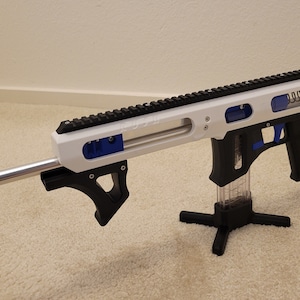 May include: A white, black, and blue toy rifle with an orange barrel extension. The rifle is supported by a black stand. The design includes a top rail, a handle, and a stock. The overall aesthetic is modern and futuristic.