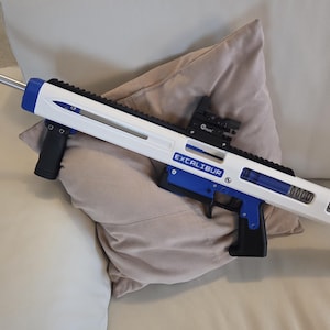 Puede incluir: Un rifle de juguete blanco y azul con punta naranja, apoyado en una almohada. El rifle tiene la palabra "EXCALIBUR" impresa en el lateral. Tiene un mango negro y una mira.