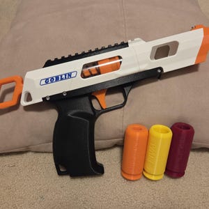 Könnte beinhalten: Eine weiß-schwarze Spielzeugpistole mit orangefarbenen und schwarzen Akzenten und dem Wort "GOBLIN" auf der Seite. Drei farbige Zylinder liegen neben der Pistole. Die Pistole liegt auf einem beigen Kissen.