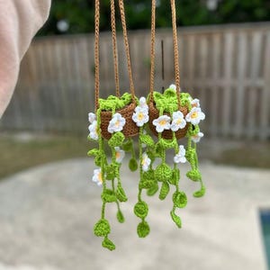 Pode incluir: Duas cestas de plantas de crochê suspensas. As cestas marrons são preenchidas com folhagem verde de crochê e flores brancas com centros amarelos. As videiras suspensas são verdes com folhas pequenas e redondas. Os cordões suspensos são marrons.