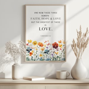 May include: A framed print with the text "AND NOW THESE THREE REMAIN: FAITH, HOPE & LOVE BUT THE GREATEST OF THESE IS LOVE." and "1 CORINTHIANS 13:13." The bottom of the print features a watercolour floral design. Two vases and a mug are on a wooden surface.