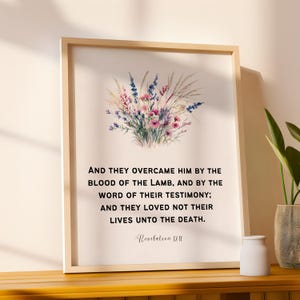 Op de afbeelding: Ingelijste print met een aquarel bloemstuk in roze, paarse en groene tinten. De print bevat de tekst "AND THEY OVERCAME HIM BY THE BLOOD OF THE LAMB... Revelation 12:11" in zwarte letters. De lijst is licht van kleur.