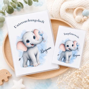 Könnte beinhalten: Zwei Baby-Aufnahmebücher aus weißem Kunststoff mit einer Illustration eines grauen Elefanten, der eine Rassel hält. Der Elefant hat rosa Ohren und ist vor einem blauen Aquarellhintergrund platziert. Der Name "Benjamin" steht auf jedem Buch.