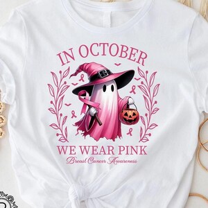May include: White t-shirt with a graphic of a pink ghost wearing a witch hat and holding a pink ribbon and a jack-o'-lantern. The text reads "IN OCTOBER WE WEAR PINK Breast Cancer Awareness".