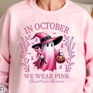 May include: A light pink sweatshirt with the text "IN OCTOBER WE WEAR PINK Breast Cancer Awareness". The graphic features a ghost wearing a witch hat and holding a pink ribbon and a jack-o'-lantern.