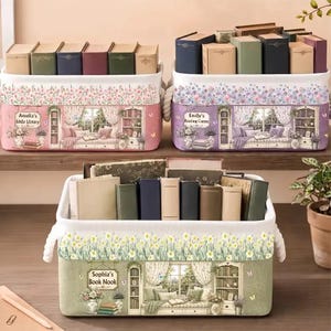 May include: Three rectangular fabric storage bins with rope handles, each filled with books. The bins feature floral patterns and printed illustrations of cozy reading nooks with text reading Amelia's Little Library, Emily's Reading Corner, and Sophia's Book Nook.