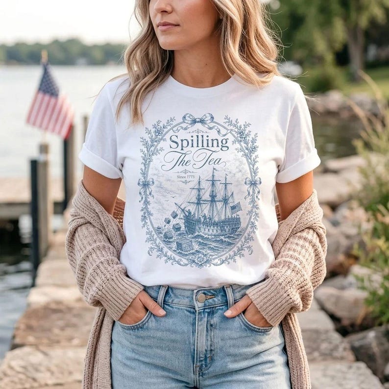 May include: A white short-sleeve t-shirt featuring a blue graphic print of a sailing ship surrounded by an ornate floral wreath. The text "Spilling The Tea" and "Since 1773" is printed in the center. The shirt is worn with a beige cardigan and blue jeans.