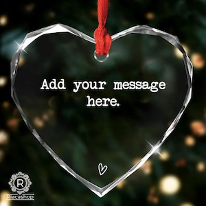 May include: A clear, heart-shaped ornament with the text "Add your message here." and a small heart symbol. It hangs from a red ribbon. The ornament is faceted, catching light, and is suitable for personalization.