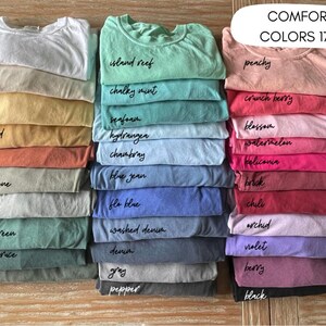 May include: A collection of folded Comfort Colors 1717 t-shirts arranged in three vertical stacks. The shirts display a wide variety of colors including white, ivory, butter, mustard, yam, sandstone, bay, light green, blue spruce, moss, island reef, chalky mint, seafoam, hydrangea, chambray, blue jean, fla blue, washed denim, denim, gray, pepper, peachy, crunch berry, blossom, watermelon, heliconia, brick, chili, orchid, violet, berry, and black.