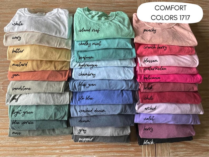 May include: A collection of folded Comfort Colors 1717 t-shirts arranged in three vertical stacks. The shirts display a wide variety of colors including white, ivory, butter, mustard, yam, sandstone, bay, light green, blue spruce, moss, island reef, chalky mint, seafoam, hydrangea, chambray, blue jean, fla blue, washed denim, denim, gray, pepper, peachy, crunch berry, blossom, watermelon, heliconia, brick, chili, orchid, violet, berry, and black.