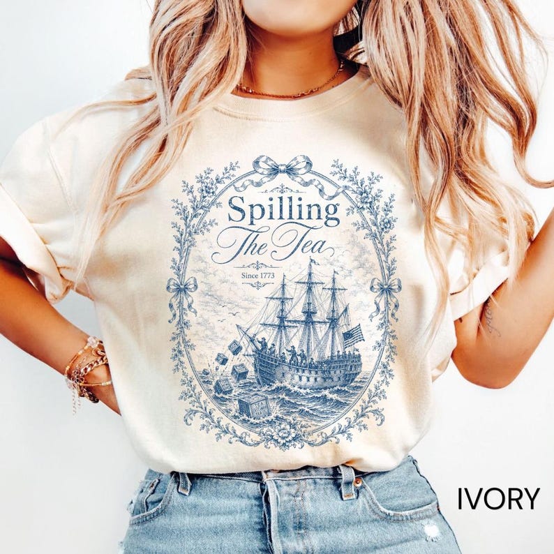 May include: An ivory t-shirt featuring a blue graphic print of a historic sailing ship tossing crates into the ocean. The design is enclosed in an ornate floral oval frame with a bow at the top and includes the text Spilling The Tea Since 1773.