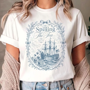 May include: A white short-sleeve t-shirt featuring a blue graphic print of a sailing ship at sea with crates falling into the water. The design is enclosed in an oval floral frame with a bow at the top and text that reads Spilling The Tea Since 1773.