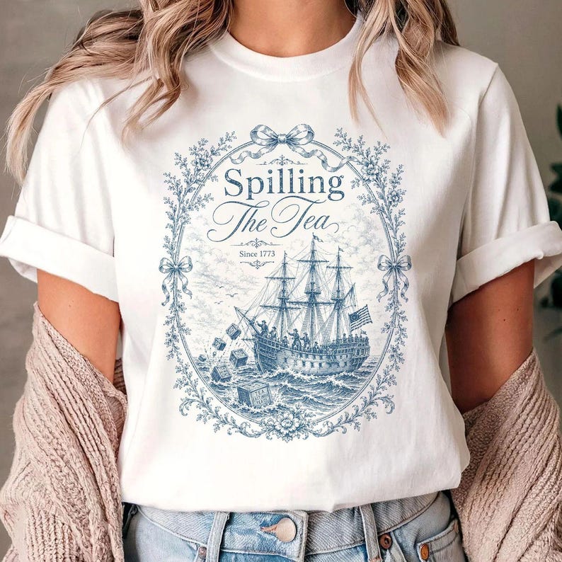 May include: A white short-sleeve t-shirt featuring a blue graphic print of a sailing ship at sea with crates falling into the water. The design is enclosed in an oval floral frame with a bow at the top and text that reads Spilling The Tea Since 1773.