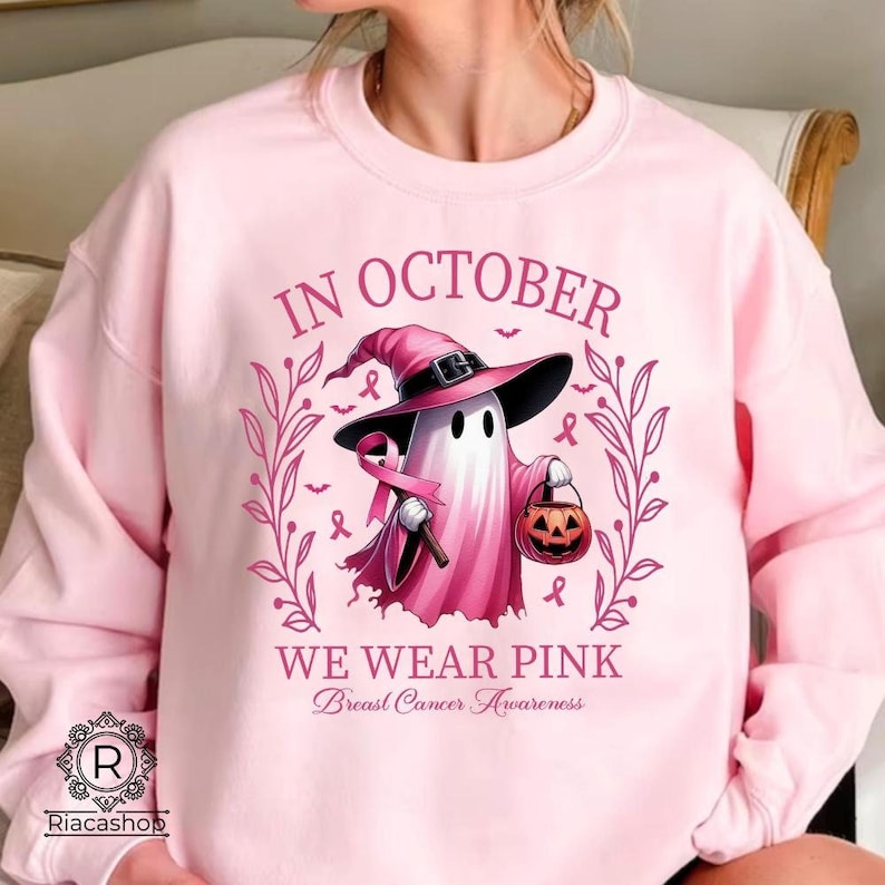 May include: A light pink sweatshirt with the text "IN OCTOBER WE WEAR PINK Breast Cancer Awareness". The graphic features a ghost wearing a witch hat and holding a pink ribbon and a jack-o'-lantern.