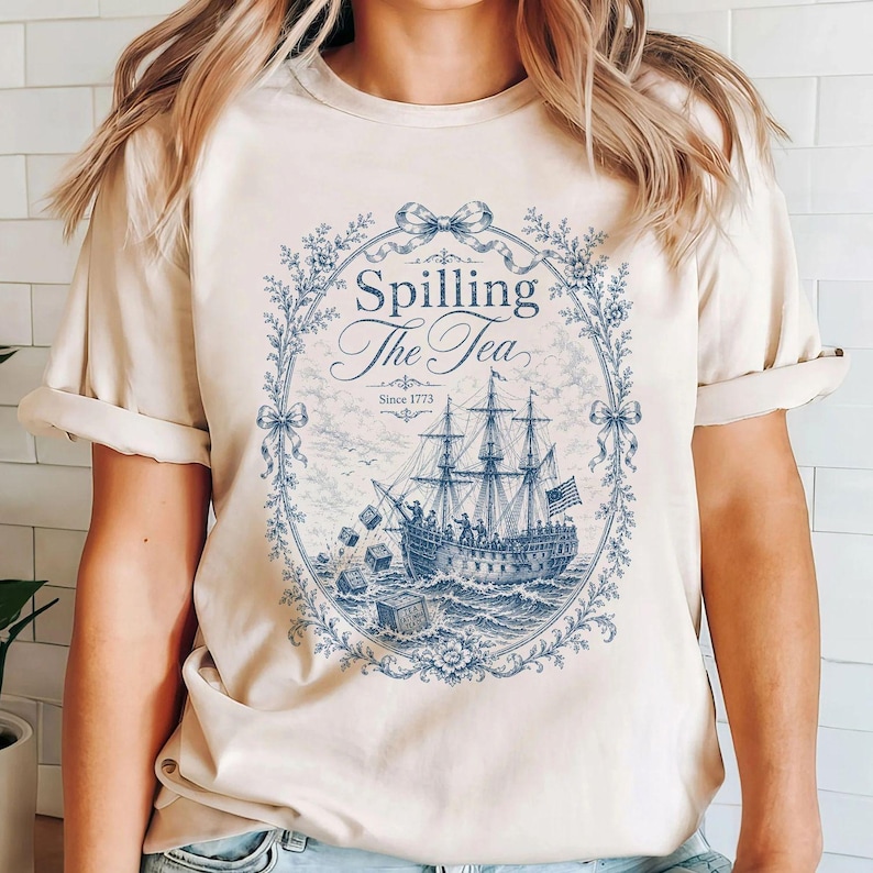 May include: A cream-colored short-sleeve t-shirt featuring a blue graphic print of a sailing ship at sea. The design includes the text "Spilling The Tea" and "Since 1773" framed by an ornate floral wreath with bows, depicting crates being tossed overboard.